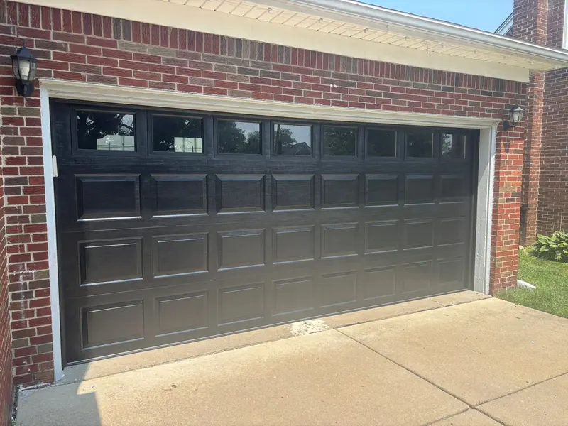 New black raised-panel double garage door with a row of windows, installed on a brick house